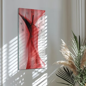 Peut inclure: Une horloge murale rectangulaire avec un motif abstrait rouge et blanc. Le cadran de l'horloge comporte des chiffres et des aiguilles noirs. L'horloge est fixée sur un mur blanc, avec de la lumière naturelle et des plantes en arrière-plan.