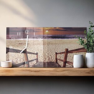 May include: A rectangular wall clock featuring a beach scene with a wooden walkway leading to the ocean at sunset. The clock face displays black numerals and hands. The clock is displayed on a wooden shelf.