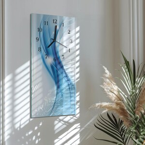 May include: A rectangular wall clock with a blue and white abstract design. The clock face features black numerals and hands. The clock is mounted on a white wall, with shadows from a window visible.