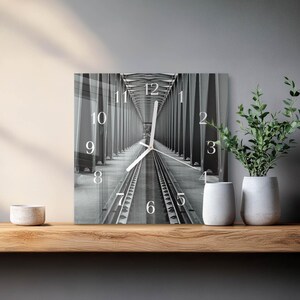 Op de afbeelding: Vierkante wandklok met een zwart-wit afbeelding van een brug en spoorrails. De wijzerplaat heeft zwarte cijfers en witte wijzers. De klok wordt getoond op een houten plank met witte vazen en groen.