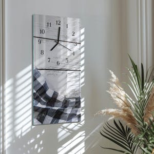 Op de afbeelding: Een rechthoekige wandklok met een witte en grijze houtlook achtergrond. De wijzerplaat heeft zwarte cijfers en wijzers. Een zwart-wit geruite deken is zichtbaar aan de onderkant. De klok is aan een witte muur bevestigd.