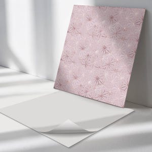 Pode incluir: Impressão retangular com um padrão geométrico de formas em ouro rosa e branco sobre um fundo rosa pálido. A impressão está encostada a uma parede branca, com uma folha de papel branca em primeiro plano.