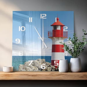 Könnte beinhalten: Eine quadratische Wanduhr mit einem Fotodruck eines rot-weißen Leuchtturms an einer felsigen Küste am Meer. Das Zifferblatt zeigt große weiße Ziffern von 1 bis 12 und dünne weiße Uhrzeiger vor der Kulisse der Küstenlandschaft.