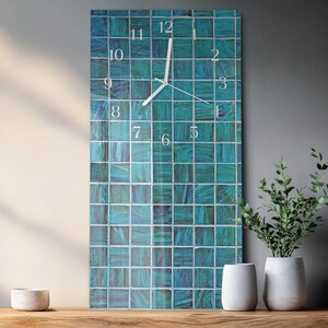 May include: A rectangular wall clock with a teal mosaic tile design. The clock face features white numerals and hands. The clock is displayed on a wooden surface, with white vases and greenery in the background.