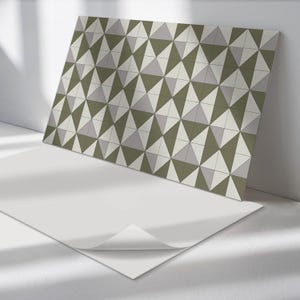 Puede incluir: Una hoja de papel tapiz autoadhesivo con un patrón geométrico de triángulos verdes, blancos y grises. El papel tapiz se muestra parcialmente despegado para revelar el respaldo adhesivo.
