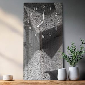 May include: A rectangular wall clock with a textured gray surface. White numerals mark the hours, and white hands indicate the time. The clock's design incorporates geometric shapes and shadows, creating a modern aesthetic. The clock is approximately 12 inches tall.