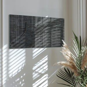 May include: A rectangular wall clock with a dark gray stone-like texture. The clock face features black hands and simple numerals. The clock is mounted on a white wall, with shadows from blinds and plants.