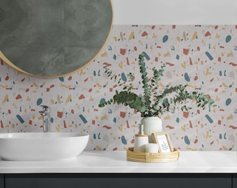 May include: A white bathroom sink with a round mirror above it. The wall behind the sink is covered in a terrazzo-style wallpaper with a white background and colorful chips. A vase with green leaves and a small tray with a soap dispenser and a picture frame are on the counter.