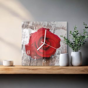 May include: Square wall clock with a large red rose design on a distressed wood-effect background. The clock has white numerals and hands. The clock is displayed on a wooden shelf with white vases and greenery.