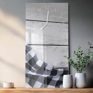 May include: A rectangular wall clock with a white face and silver hands, set against a backdrop of white-painted wooden planks. A blue and white checkered cloth drapes over the bottom of the clock. Decorative plants and vases are on a wooden surface.