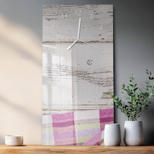 Op de afbeelding: Een rechthoekige wandklok met een witte houten achtergrond en een heldere glazen voorkant. De klok heeft witte wijzers en cijfers. De onderkant van de klok heeft een roze, paars en groen geruit patroon.