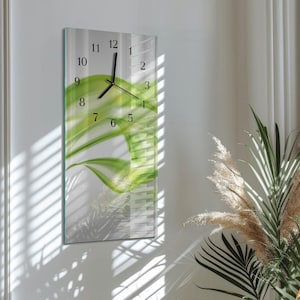 May include: A rectangular wall clock with a green and white floral design. The clock face features black numerals and hands. The clock is mounted on a white wall, with sunlight casting shadows.