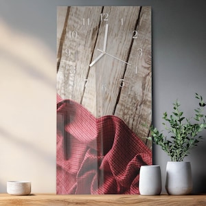 May include: A rectangular wall clock with a wooden plank background and white numerals. The clock hands point to approximately 1:05. A red patterned fabric drapes across the bottom. The clock is displayed on a wooden shelf with decorative vases and plants.