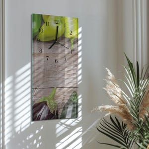 Op de afbeelding: Een rechthoekige wandklok met een groentethema. De wijzerplaat heeft een houten achtergrond met afbeeldingen van groene paprika's en aubergines. Zwarte wijzers geven de tijd aan. De klok is aan een witte muur bevestigd.