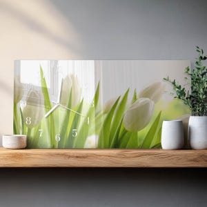 May include: Rectangular wall clock featuring a floral design with white tulips and green leaves. The clock face has white numerals and hands. The clock is displayed on a wooden shelf with decorative vases and plants.