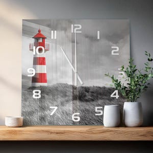 Könnte beinhalten: Eine quadratische Wanduhr mit einem Schwarz-Weiß-Foto einer grasbewachsenen Dünenlandschaft und einem rot-weiß gestreiften Leuchtturm. Das Zifferblatt zeigt große weiße Ziffern von 1 bis 12 und zwei dünne silberne Zeiger in der Mitte.