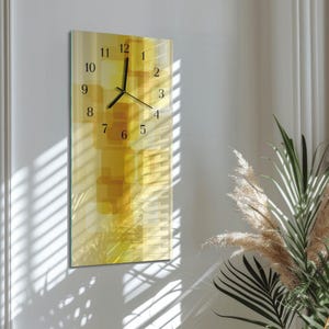 May include: A rectangular wall clock with a yellow and gold abstract design. The clock face has black numerals and hands. The clock is mounted on a white wall, with shadows from a window and plants in the corner.