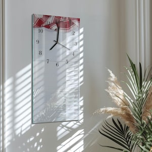 Op de afbeelding: Een rechthoekige glazen wandklok met een rood-wit geruit patroon aan de bovenkant. De wijzerplaat heeft zwarte cijfers en wijzers. De klok is aan een witte muur bevestigd.