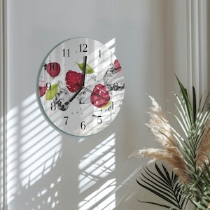 May include: A round wall clock with a white face, featuring red raspberries, green leaves, and ice cubes. Black numerals mark the hours. The clock has a clear glass cover and a light green border. The clock is hanging on a white wall.