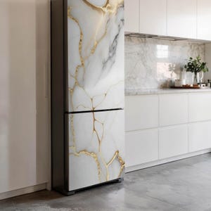 Può includere: Un frigorifero con un design in marmo bianco e venature dorate. L'elettrodomestico a due ante ha una cornice grigio scuro ed è posto contro una parete e una cucina bianche. Il motivo marmoreo aggiunge un tocco di lusso alla cucina.
