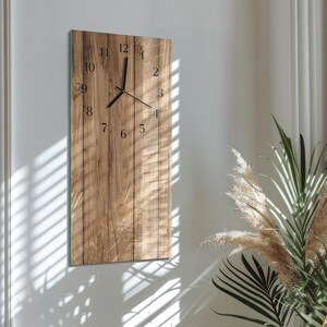 Puede incluir: Un reloj de pared rectangular de madera con un acabado de vetas de madera natural. La esfera del reloj tiene números y manecillas negras. El reloj está montado en una pared blanca, con una planta en la esquina.