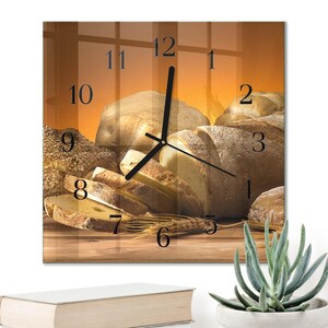 Puede incluir: Reloj de pared cuadrado con un diseño temático de pan. La esfera del reloj muestra una naturaleza muerta de varios panes, incluyendo hogazas rebanadas y enteras, con espigas de trigo. Las manecillas del reloj son negras.