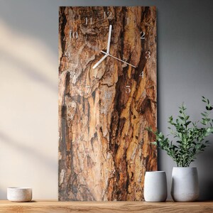 Op de afbeelding: Een rechthoekige wandklok met een natuurlijk boomschorsontwerp. De wijzerplaat van de klok heeft witte wijzers en zilveren cijfers. De klok wordt weergegeven op een houten oppervlak, met witte vazen en groen in de achtergrond.