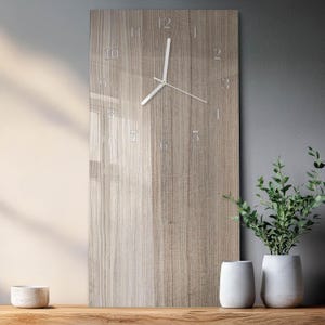 May include: A rectangular wall clock with a light wood grain finish. The clock face features silver numerals and hands. The clock is mounted on a gray wall, with a small white vase and two white planters with greenery in the foreground.