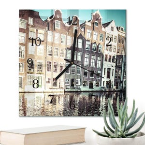 Könnte beinhalten: Quadratische Wanduhr mit einer malerischen Szene von Grachtenhäusern in Amsterdam. Das Zifferblatt zeigt große schwarze Ziffern und schwarze Zeiger. Das Bild zeigt die Spiegelung der Gebäude im Wasser. Ein dekorativer Einrichtungsgegenstand.
