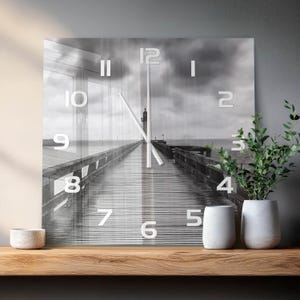 Könnte beinhalten: Eine quadratische Wanduhr mit einer Schwarz-Weiß-Fotografie eines Holzstegs, der unter einem bewölkten Himmel auf einen Leuchtturm zuführt. Das Zifferblatt zeigt weiße arabische Ziffern und zwei dünne, mittig platzierte weiße Zeiger.