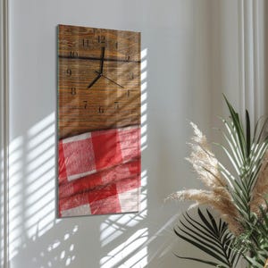 Op de afbeelding: Een rechthoekige wandklok met een houtnerf ontwerp en zwarte wijzers. De onderste helft van de klok heeft een rood-wit geblokt patroon. De klok is aan een witte muur bevestigd.