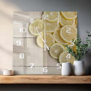 May include: A square wall clock featuring a photographic print of sliced yellow lemons layered over a light-colored wooden plank background. The clock face displays white Arabic numerals from 1 to 12 and two thin white clock hands centered.