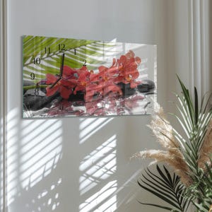 May include: Rectangular wall clock featuring a serene image of red orchids, black stones, and green palm leaves. The clock face displays black hands and numerals against the floral backdrop. The clock is mounted on a white wall.
