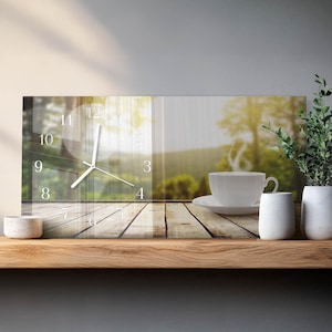 May include: A rectangular wall clock with a transparent face, displaying the time. A white teacup and saucer sit on a wooden surface, with a blurred outdoor scene in the background. Decorative white vases and a small bowl are also present.