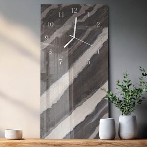 May include: A rectangular wall clock with a marble-like design in shades of gray and white. The clock face features silver numerals and hands. The clock is displayed on a wooden surface, with decorative plants in white pots.