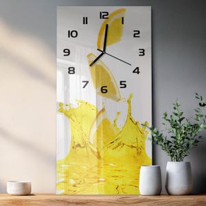 Può includere: Un orologio da parete rettangolare con una stampa grafica di tre fette di limone giallo che cadono in uno spruzzo di liquido giallo. Il quadrante mostra numeri arabi neri da 1 a 12 e lancette nere per ore, minuti e secondi.