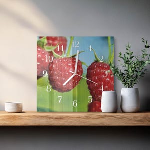 May include: Square wall clock featuring a close-up of ripe red raspberries. The clock face displays white numerals and hands against a backdrop of the fruit and green leaves. The clock is on a wooden shelf.