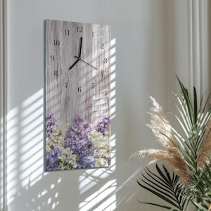 Könnte beinhalten: Eine rechteckige Wanduhr mit Holzoptik und einem Blumendesign am unteren Rand. Das Zifferblatt hat schwarze Zahlen und Zeiger. Das Blumendesign zeigt weiße, violette und lavendelfarbene Blumen.