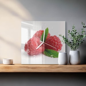 May include: Square wall clock with a close-up of red raspberries and green leaves. The clock face has white numerals and hands. The clock is displayed on a wooden shelf with white vases and a plant.