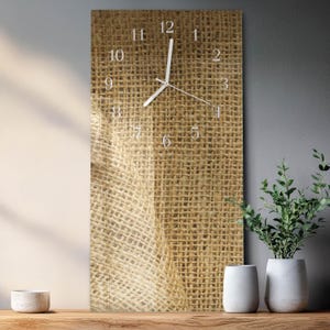 May include: A rectangular wall clock with a burlap texture background. The clock face features white numerals and hands. The clock is displayed on a wooden surface, with white vases and greenery in the background.