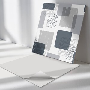 May include: Abstract art print featuring a pattern of gray and blue geometric shapes on a white background. The design includes squares, rectangles, and brushstroke-like elements. The artwork is displayed against a white wall.