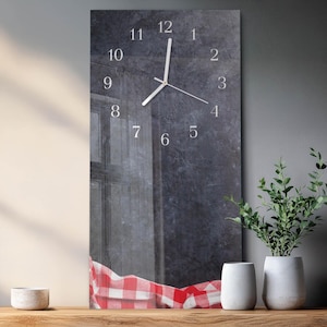 May include: A rectangular wall clock with a dark gray textured background and white numerals. The clock hands point to 12:07. A red and white checkered tablecloth is visible at the bottom. Two white vases and a potted plant are on a wooden surface.