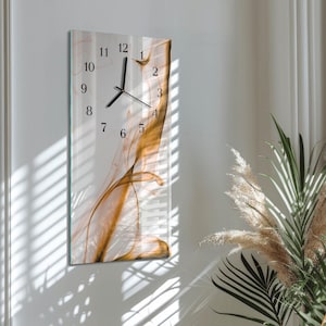 Può includere: Orologio da parete rettangolare con quadrante in vetro. L'orologio presenta numeri e lancette neri su sfondo bianco con un design astratto e fluido nei toni dell'oro e del marrone. L'orologio è montato su una parete bianca.