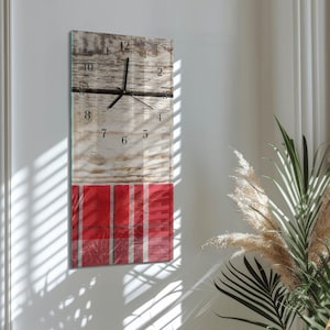 Op de afbeelding: Een rechthoekige wandklok met een houtnerf ontwerp en een rood geruit patroon aan de onderkant. De klok heeft zwarte cijfers en wijzers. De klok is aan een witte muur bevestigd.
