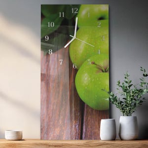 May include: A rectangular wall clock featuring a close-up of green apples on a wooden surface. The clock face has silver numerals and hands. The background shows more green apples and leaves. The clock is displayed on a wooden shelf.