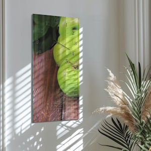 Op de afbeelding: Een rechthoekige wandklok met een afbeelding van groene appels op een houten oppervlak. De wijzerplaat van de klok toont zwarte cijfers en wijzers, met de nummers 1 tot en met 12. De klok is aan een witte muur bevestigd.