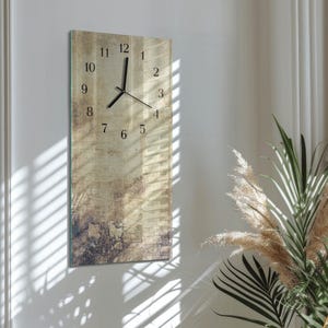 Puede incluir: Un reloj de pared rectangular con un fondo texturizado beige y marrón. Los números negros marcan las horas y las manecillas negras indican la hora. El reloj está montado en una pared blanca, con sombras de persianas visibles.
