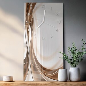 Op de afbeelding: Een rechthoekige wandklok met een modern design. De wijzerplaat heeft een lichte achtergrond met bruine en beige abstracte golfpatronen. Zilverkleurige wijzers en cijfers geven de tijd aan. De klok wordt tentoongesteld met decoratieve vazen ​​en planten.