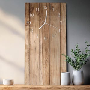 May include: A rectangular wall clock with a wood grain design. The clock face features white numerals and hands. The clock is displayed on a wooden surface, with two white vases and a small bowl. The background is a neutral gray.