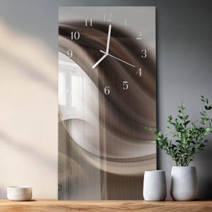 May include: A rectangular wall clock with a brown and beige abstract design. The clock face has white numerals and hands. The clock is displayed on a wooden surface with two white vases and a potted plant.